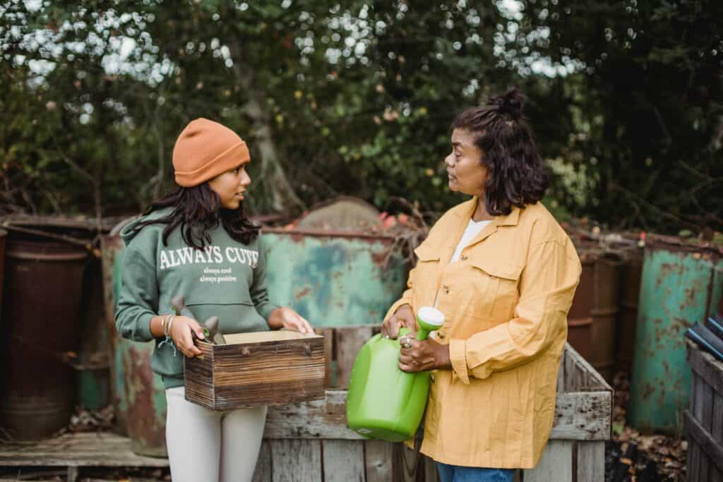 Adult woman standing holding a green watering can talking to a young person holding a gardening box