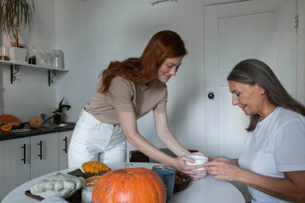 Two women in a kitchen. One woman is standing and is handing the other woman who is seated a cup of tea on a table.